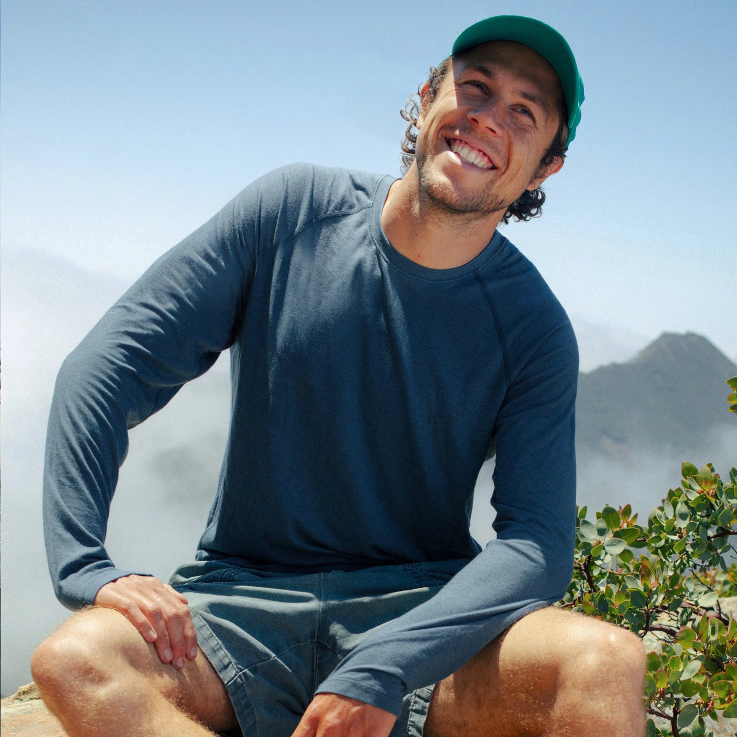 Three-quarter front view of Men’s Sol Crew long-sleeve UPF sun shirt in deep blue on a smiling model showing relaxed fit with ocean and rocky cliffs background