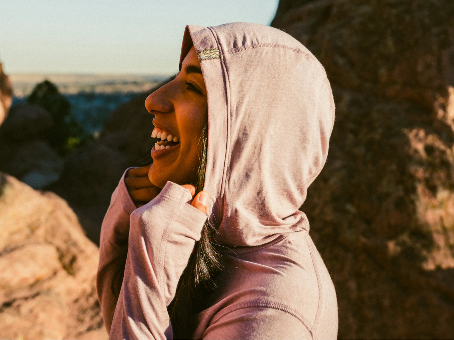 Model smiling in profile pulling hood up wearing light pink alpaca long-sleeve UPF shirt in regular fit, rocky outdoors
