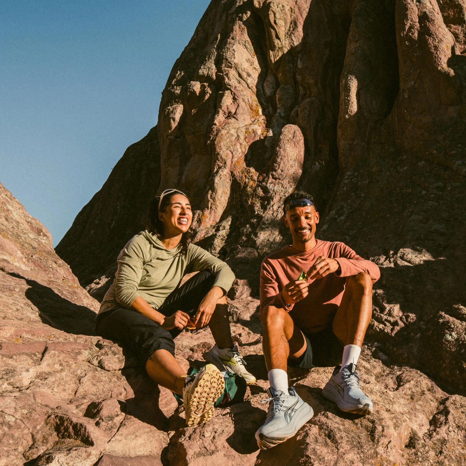 Two hikers smiling and resting on sunlit rocks wearing long-sleeve UPF sun shirts (man rust, woman olive)