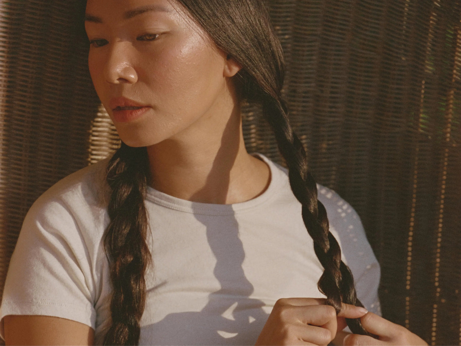 Woman adjusting her braided hair while wearing a white crew-neck tee against a woven wicker background