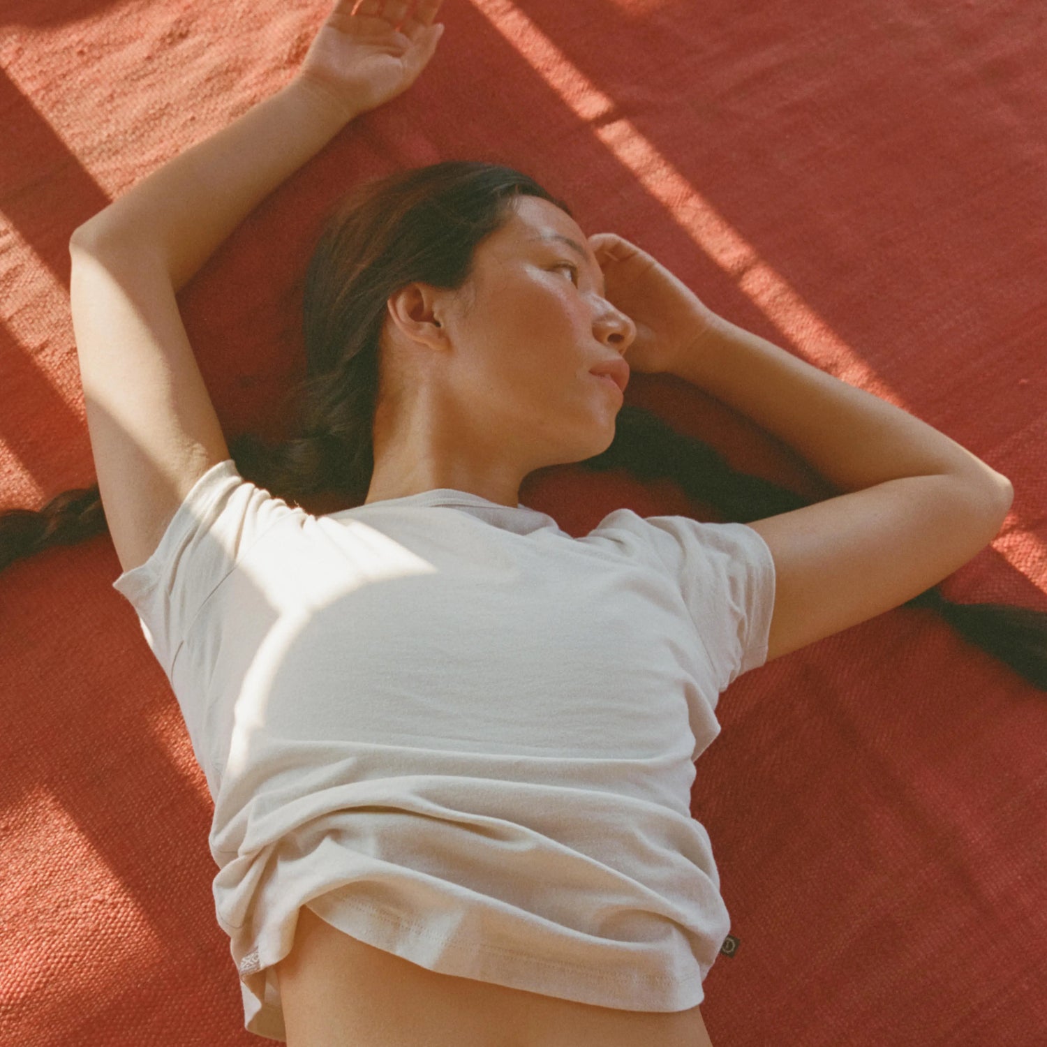 Woman lying on a red rug wearing an off-white Women’s Essential Tee in a relaxed, sunlit pose
