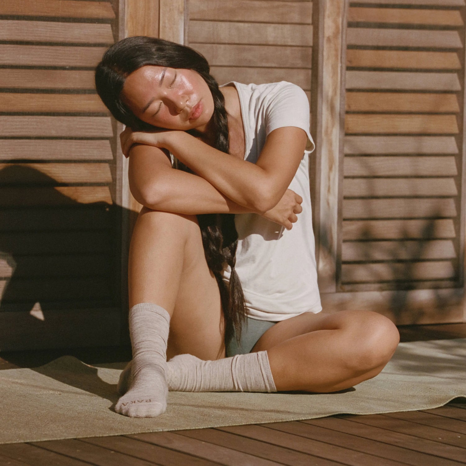 Woman wearing a white V-neck tee in a relaxed fit, seated hugging her knees against wooden shutters in warm sunlight