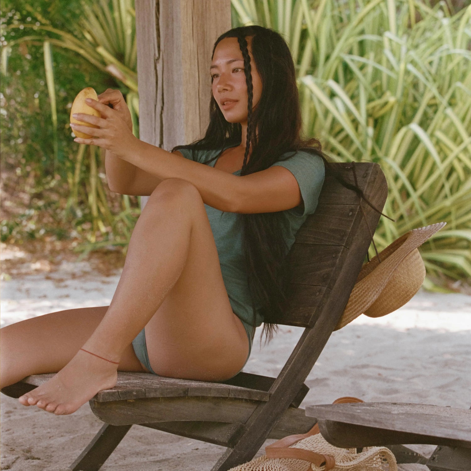 Woman wearing teal V-neck tee, sitting on a wooden beach chair and holding a mango with tropical plants behind her