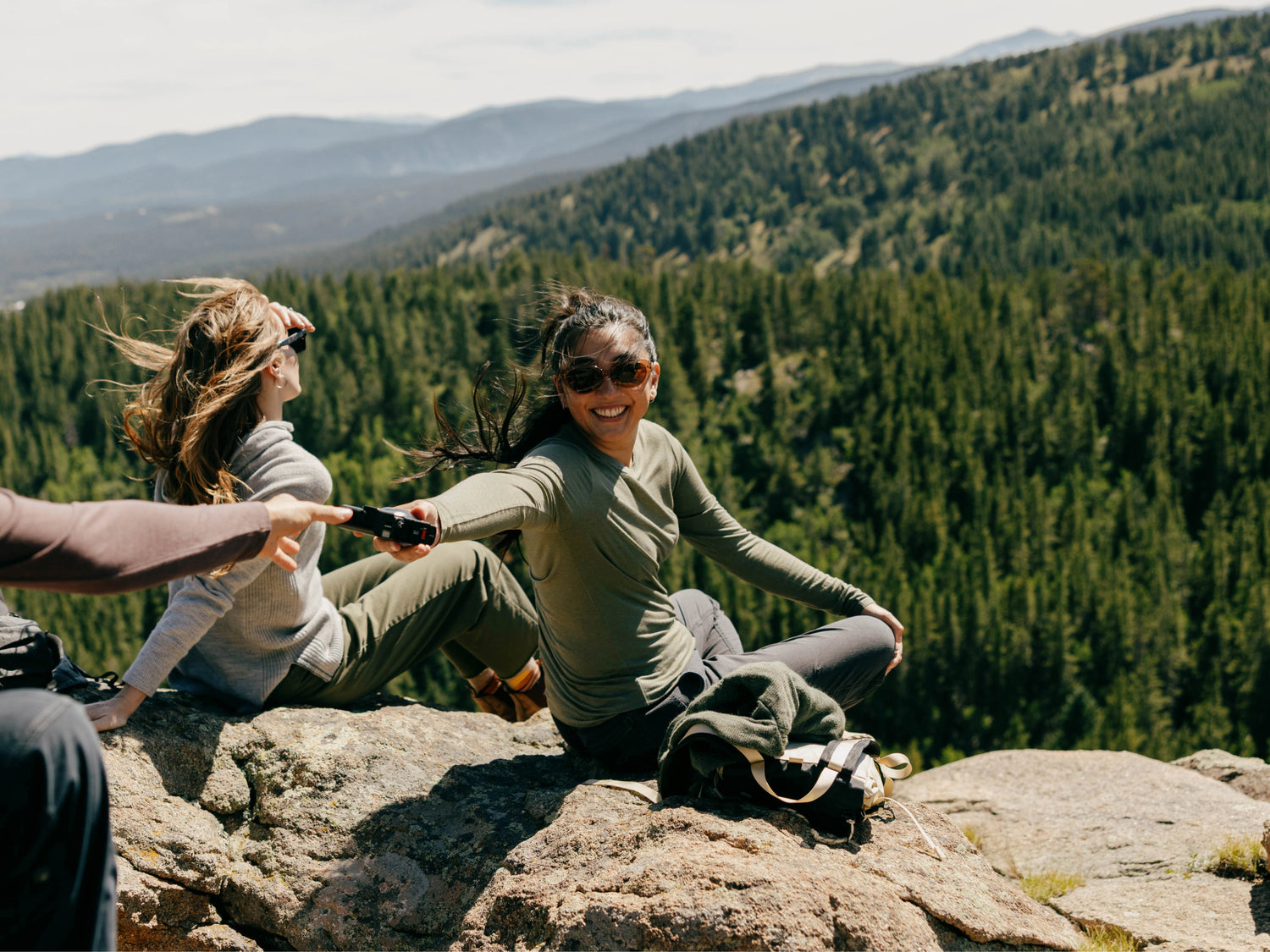 Woman in olive Women’s Aire long sleeve shirt passing a device while seated on rocks with forested mountains behind