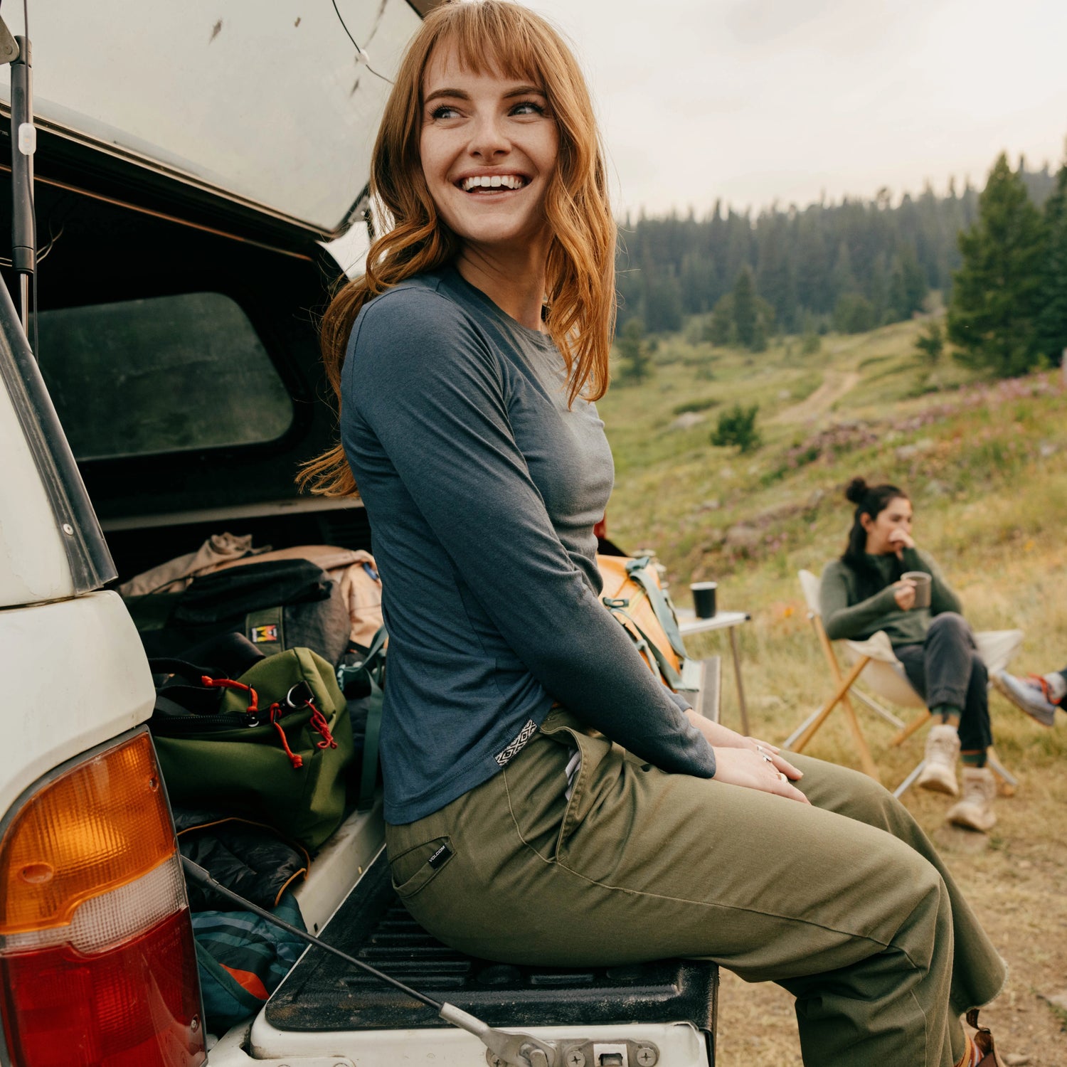 Woman sitting on a truck tailgate wearing a slate blue Women’s Aire Long Sleeve slim fit with camping gear at a grassy mountain campsite