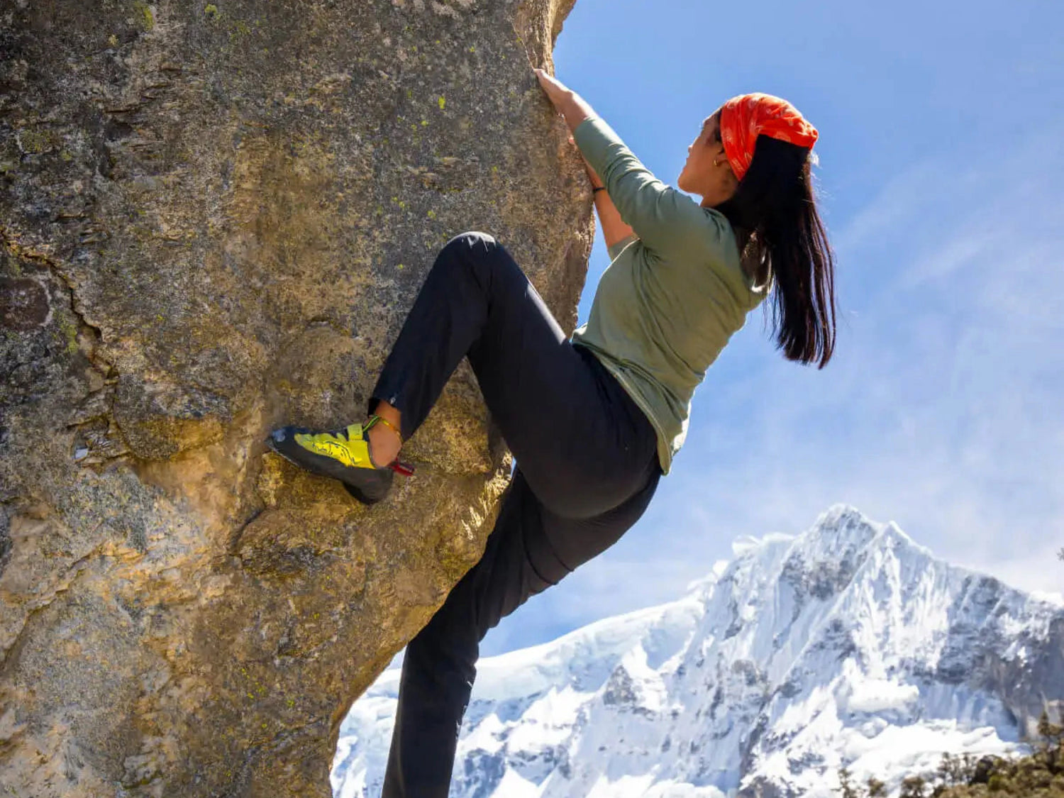 Woman wearing olive Women’s Aire Long Sleeve slim fit climbing a boulder with snowy mountain peaks in the background