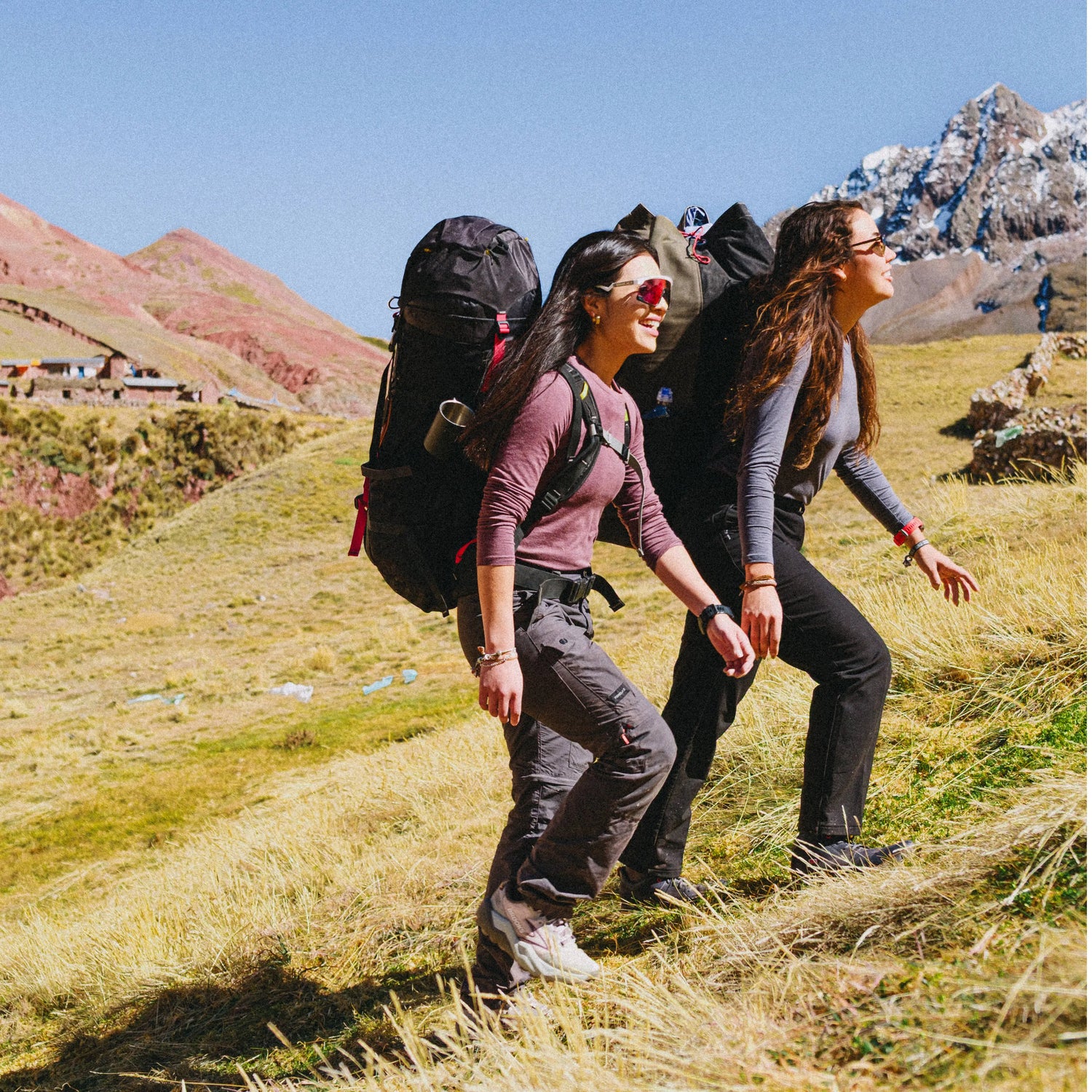 Two women hiking in a mountain meadow wearing Women’s Aire Long Sleeve shirts, one burgundy and one gray, carrying backpacks