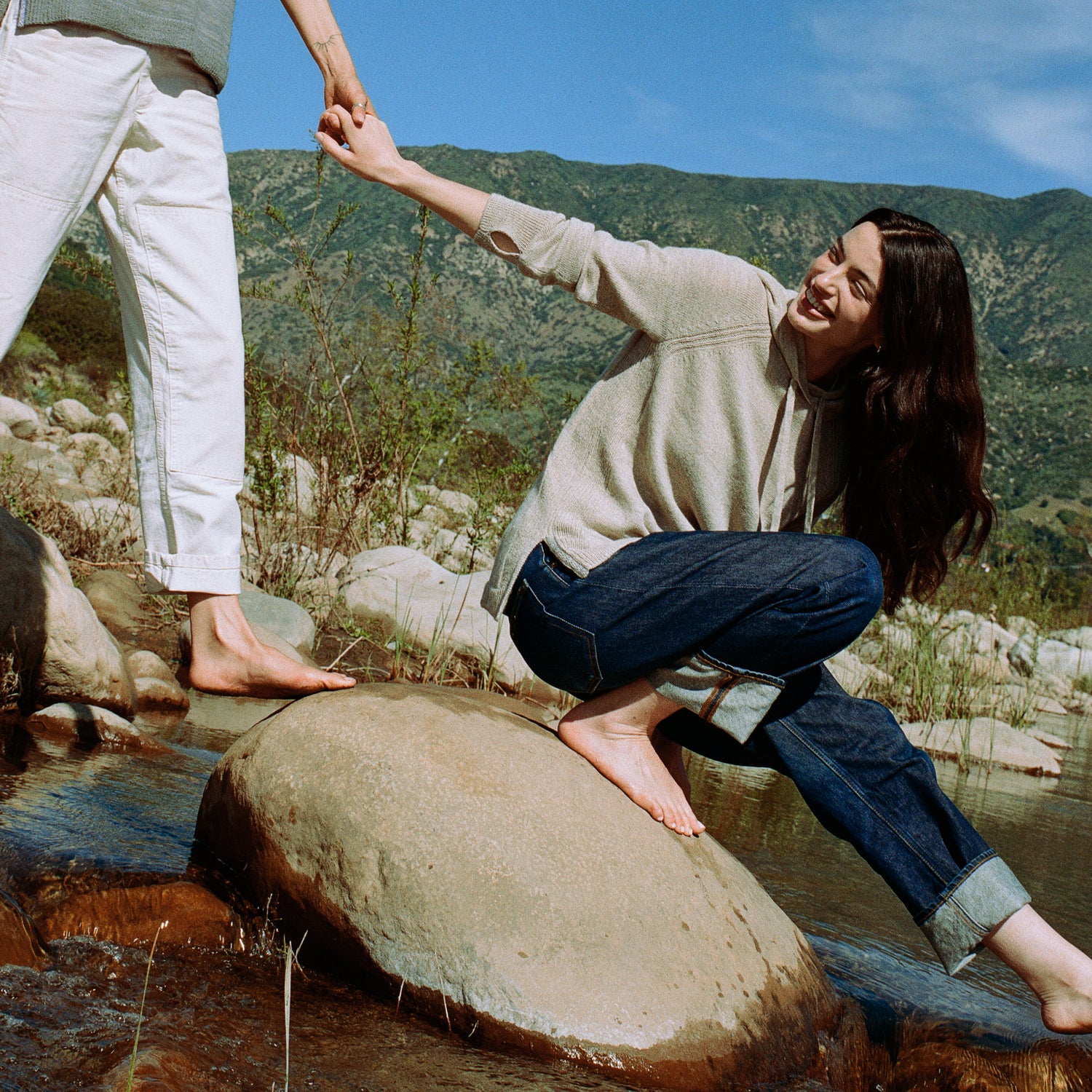 Woman wearing a beige alpaca hoodie kneeling on rocks and holding another person’s hand beside a mountain stream
