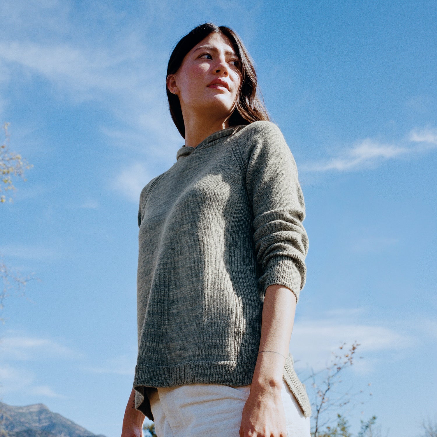 Low angle view of women’s alpaca hoodie in spruce green, relaxed fit showing ribbed cuffs and hood against blue sky