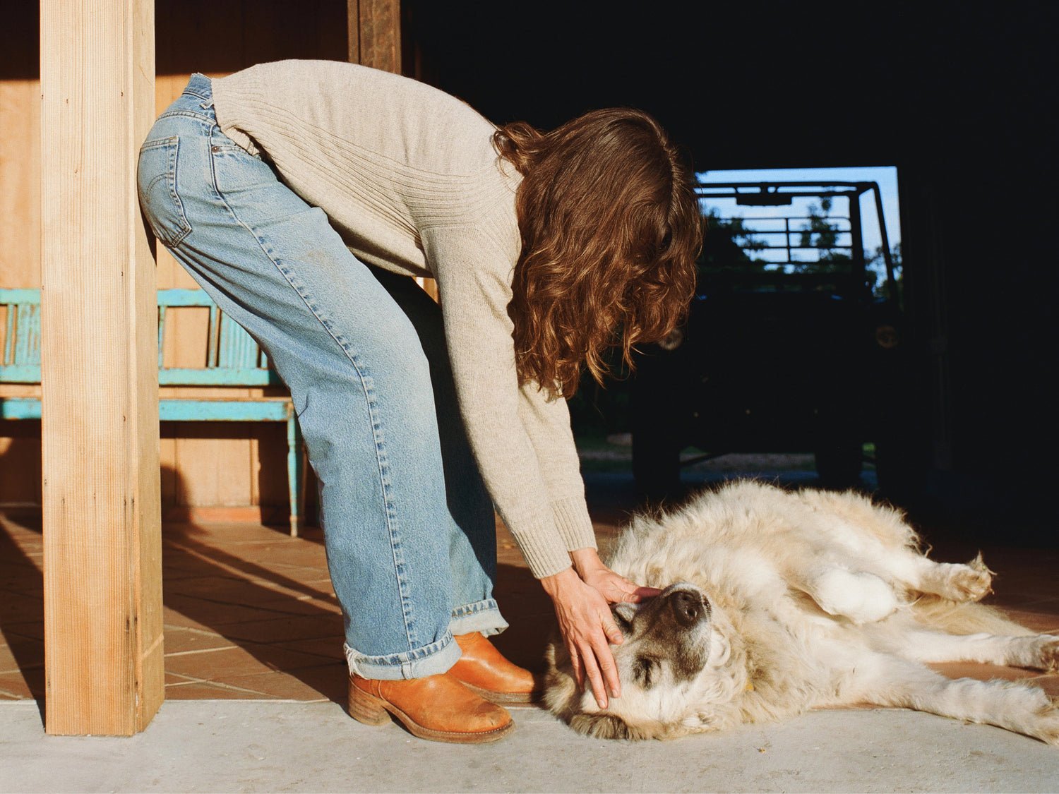 Woman bending to pet a large dog on a sunlit porch, wearing a light beige women’s alpaca crewneck sweater in relaxed fit