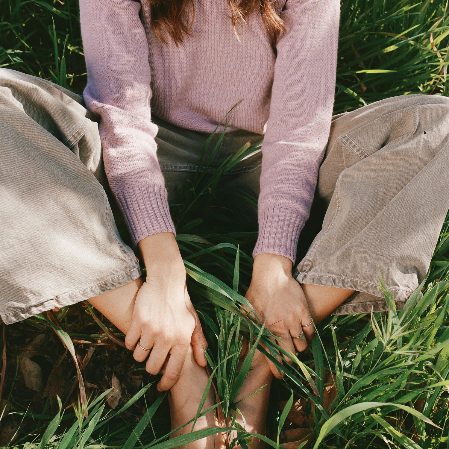 Woman sitting cross-legged wearing a dusty pink women’s alpaca crewneck sweater in a relaxed fit, seated in tall green grass