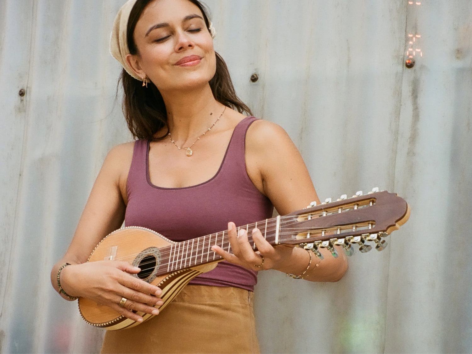 Woman playing a guitar against a corrugated metal wall, wearing a mauve tank top and headscarf