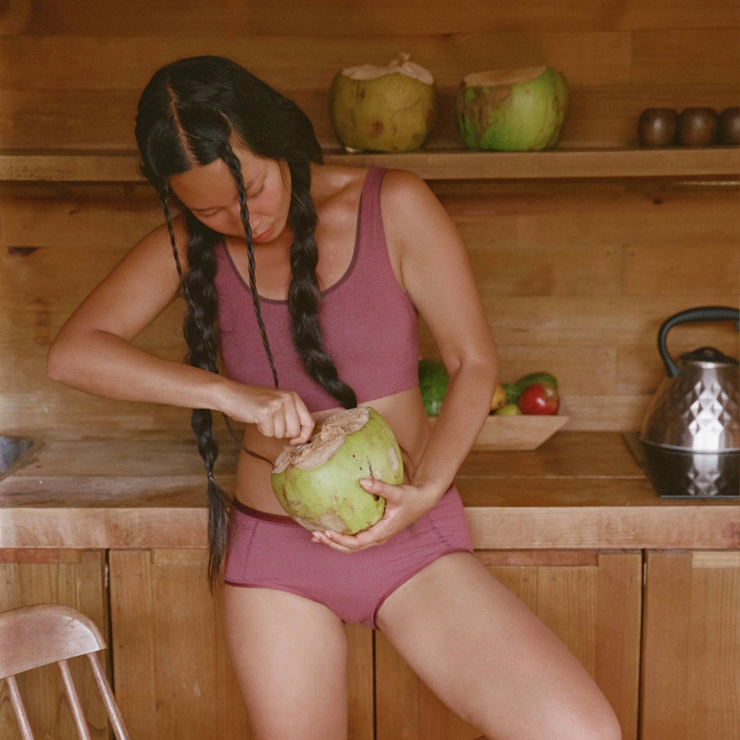 Woman opening a coconut while wearing a mauve scoop bralette and matching briefs in a wooden kitchen setting