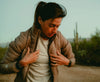 Woman zipping a tan Mayu ultralight insulated jacket outdoors in a desert-like landscape