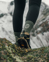Hiker standing on a rocky mountain trail wearing olive-green patterned performance crew socks