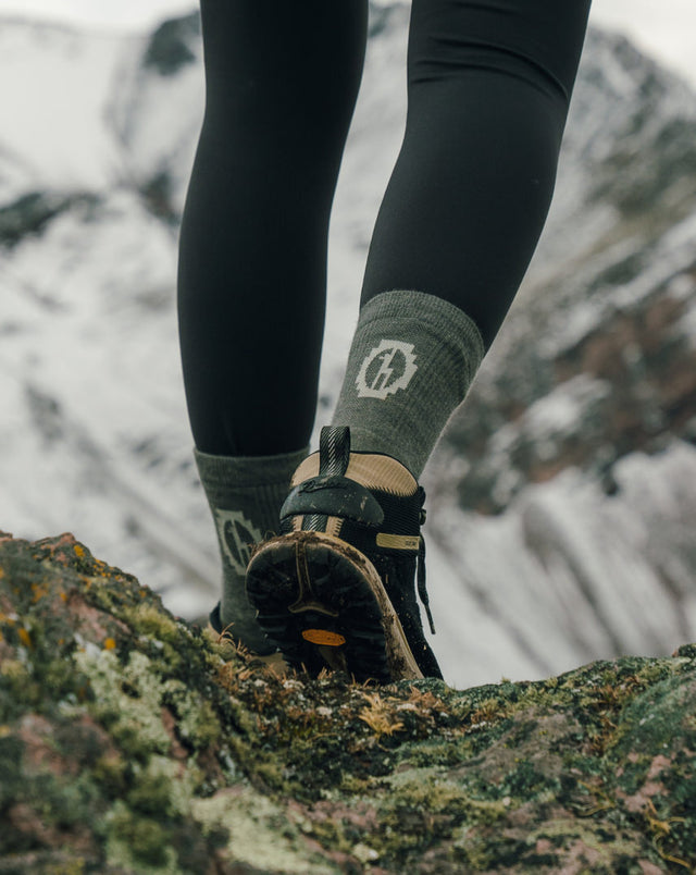 Hiker standing on a rocky mountain trail wearing olive-green patterned performance crew socks