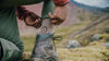 Hiker kneeling to adjust a shoe on a rocky mountain trail with another hiker and rolling hills in the background