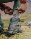 Hiker pulling on a tan performance crew sock inside a worn hiking boot on a mossy mountain trail