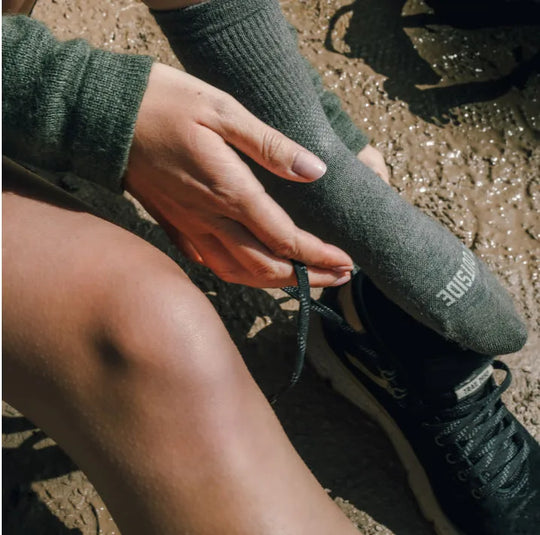 Hiker adjusting a black crew sock on their lower leg while sitting on a rocky trail