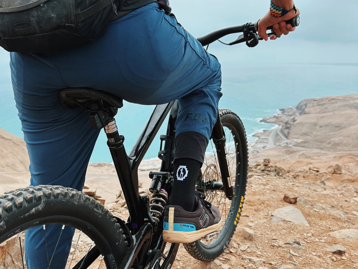 Rear view of Trail 3/4 Crew socks showcasing black three-quarter length, white logo, and snug fit on a mountain biker at a rocky coastal overlook