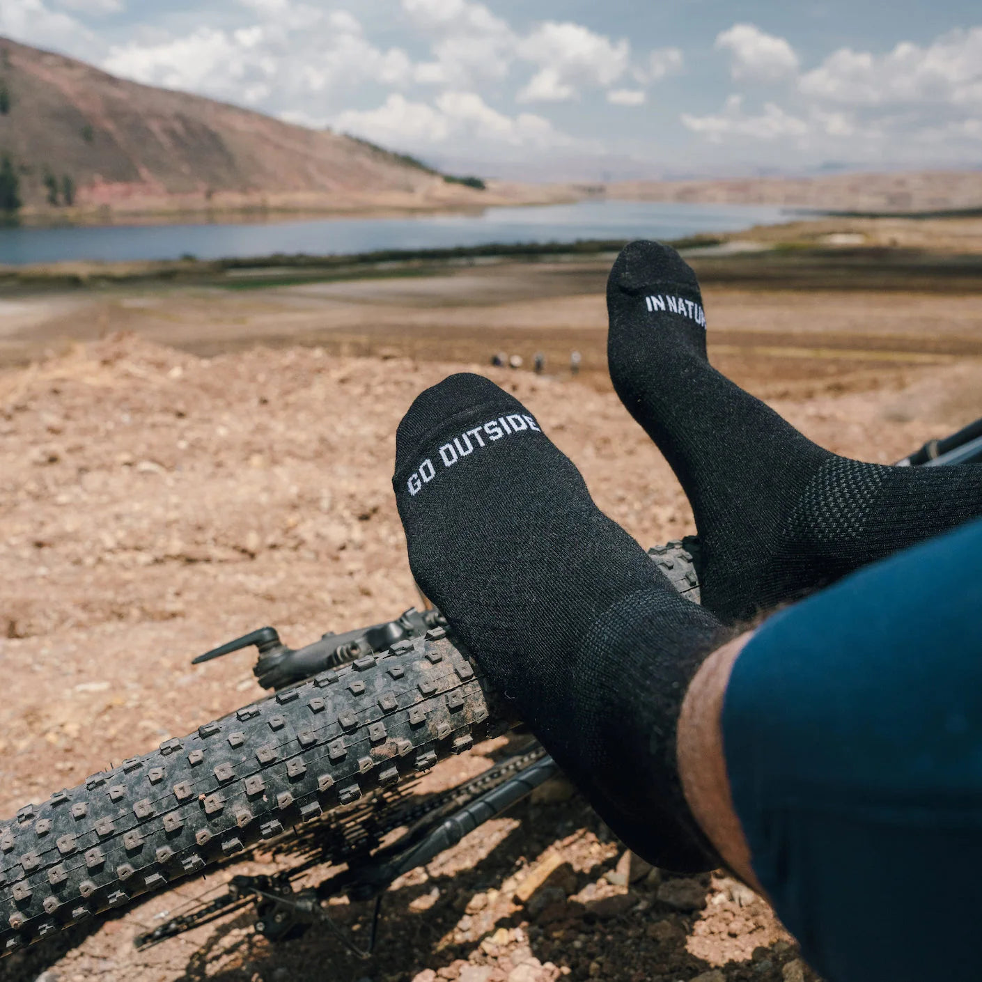 Feet resting on a mountain bike tire wearing black 3/4 crew socks that read ‘GO OUTSIDE’ and ‘IN NATURE’ with a lakeshore background