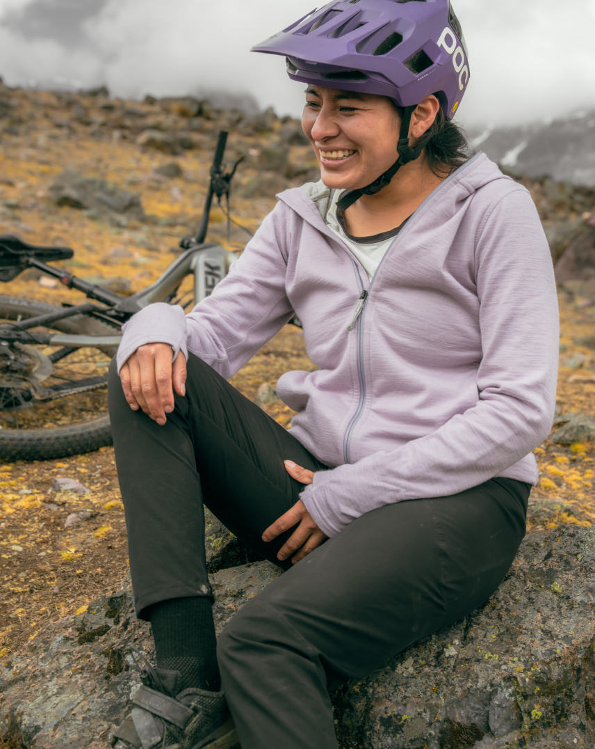 Smiling cyclist sitting on a rock wearing black Trail Crew hiking socks, purple helmet and mountain bike in misty alpine