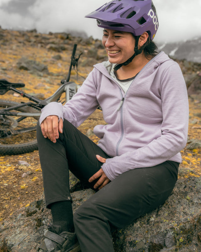 Smiling cyclist sitting on a rock wearing black Trail Crew hiking socks, purple helmet and mountain bike in misty alpine