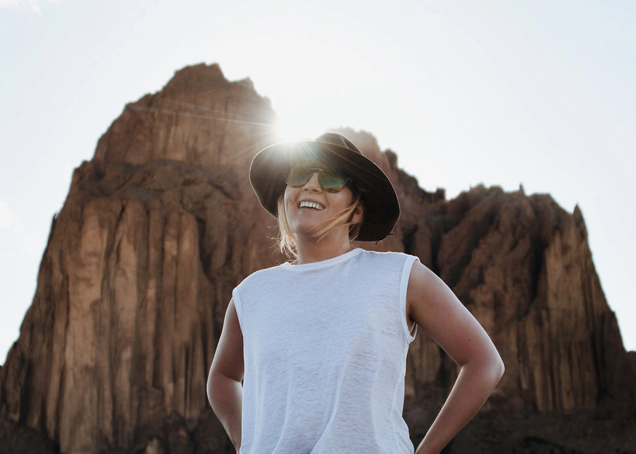 Person wearing a white sleeveless shirt and black hat with sunglasses, standing in front of desert rock formations.