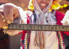 Quechua weaver holding a colorful handwoven bracelet band toward the camera with Pisac village blurred behind