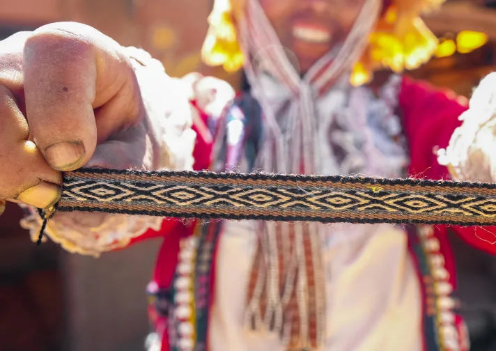 Quechua weaver holding a colorful handwoven bracelet band toward the camera with Pisac village blurred behind