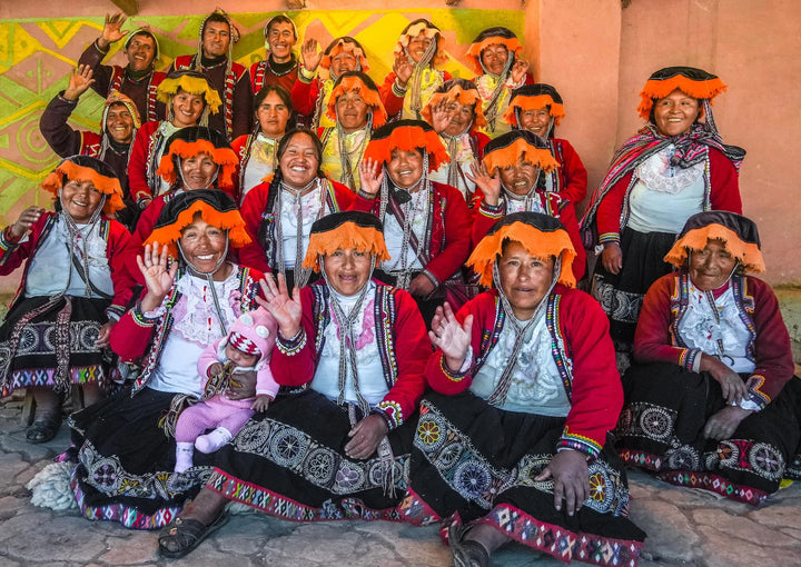 Quechua women weavers smiling and waving outside Pisac wearing traditional red jackets, embroidered skirts, and orange headpieces
