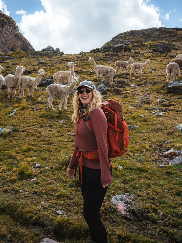 Hiker in a red jacket and backpack standing among grazing llamas on a grassy Andean slope with rocky peaks behind