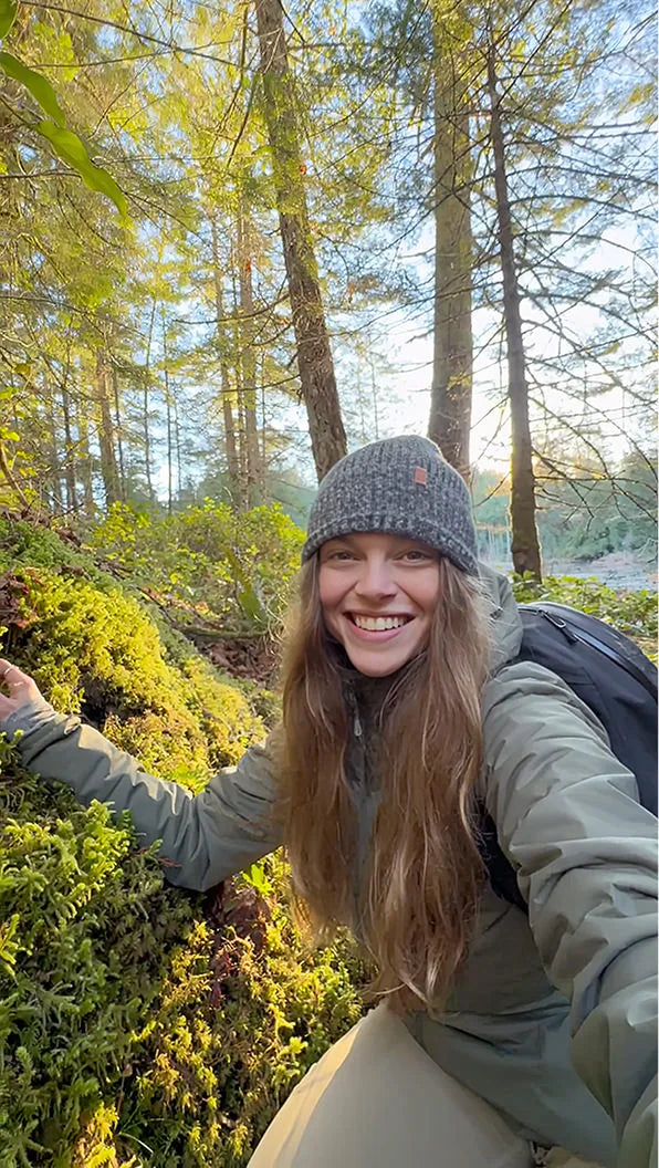 Rachael Tancock smiling in a sunlit forest, crouching on moss with a green jacket, gray knit beanie, and backpack