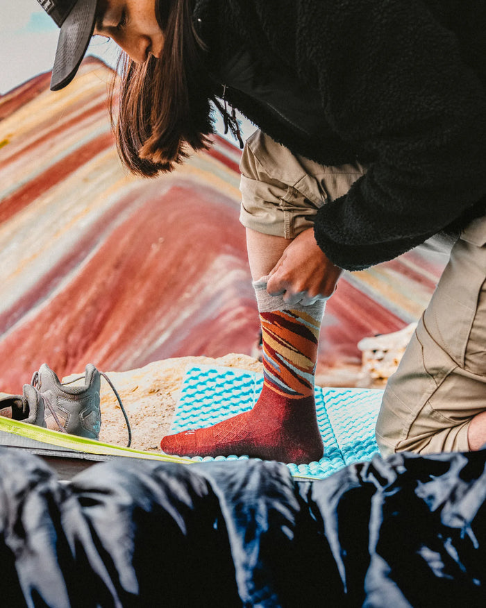 Hiker pulling on a Rainbow Mountain crew sock while kneeling on a blue foam mat with colorful striped mountain in the background