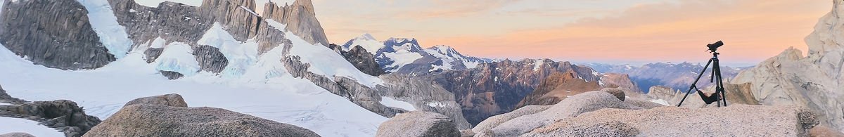 Tripod-mounted camera on a rocky ridge overlooking snow-covered mountains and glaciers at sunrise