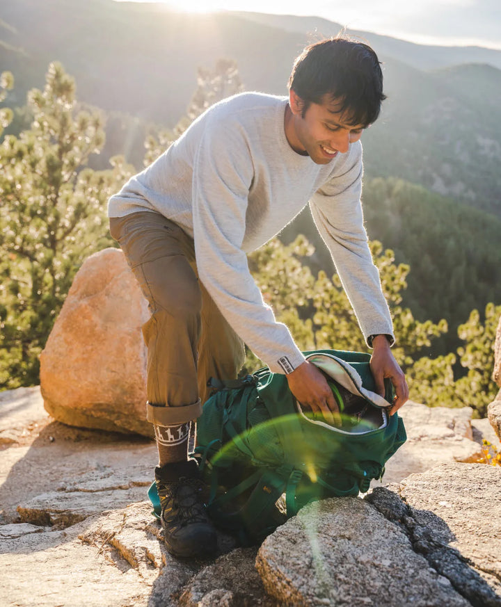 A smiling man in the nature getting some thing from his backpack. He's wearing an alpaca sweater and our brown costa socks. 