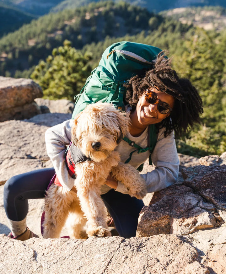 A smiling backpacker girl smiling outdoors. She's on her knees hugging her cute dog 