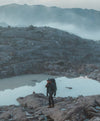 Hiker walking across a rocky volcanic landscape with pools of water and misty mountains in the background