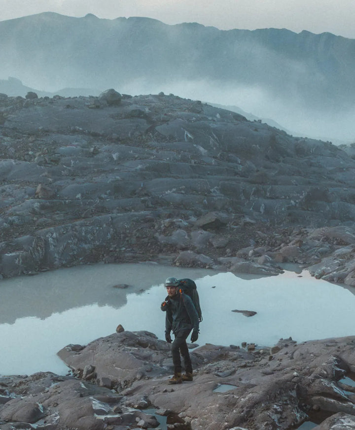 Hiker walking across a rocky volcanic landscape with pools of water and misty mountains in the background