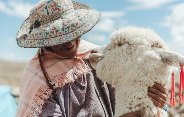 Person standing with an alpaca in a grassy field, wearing a straw hat and knit sweater