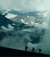 Three climbers hiking along a snowy Andean ridge with towering peaks and low clouds in the background