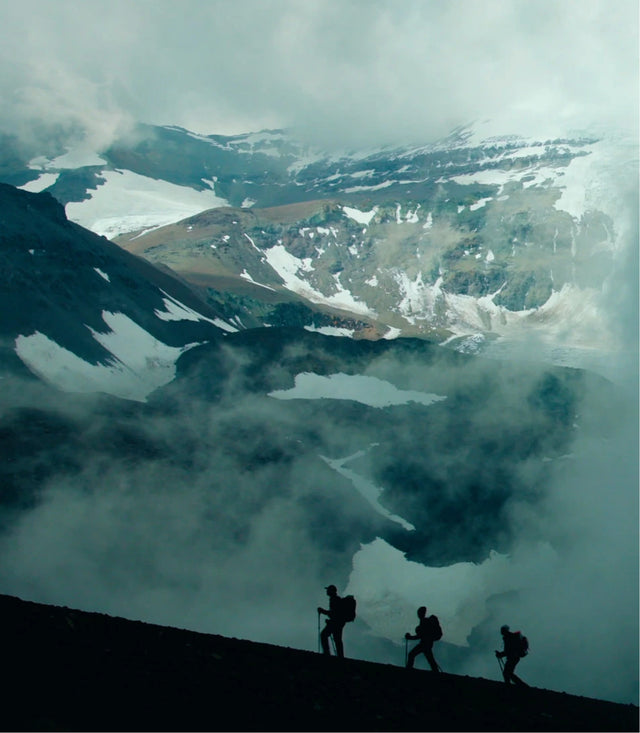 Three climbers hiking along a snowy Andean ridge with towering peaks and low clouds in the background