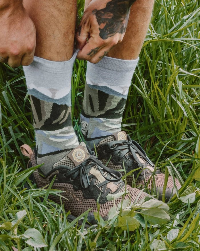 Person pulling on light gray crew alpaca socks with green and blue mountain graphic while standing in grass wearing hiking shoes