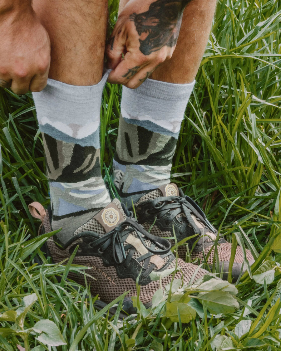 Person pulling on light gray crew alpaca socks with green and blue mountain graphic while standing in grass wearing hiking shoes