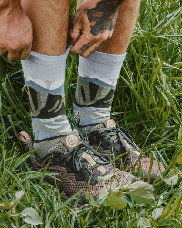 Person pulling on light gray crew alpaca socks with green and blue mountain graphic while standing in grass wearing hiking shoes