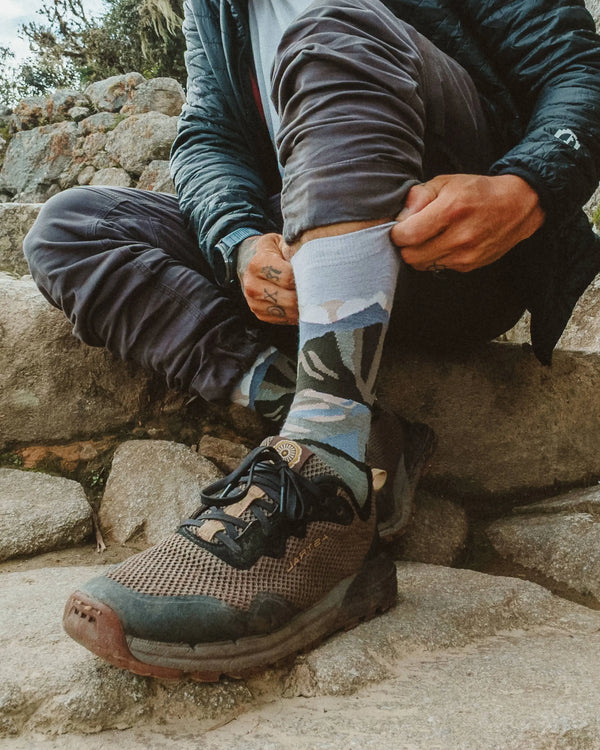 Person pulling up light blue mountain-pattern alpaca crew socks while sitting on rocks during a hike