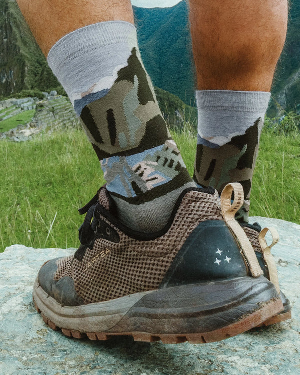 Close-up of Essential Sacred Valley crew socks showing mountain graphic and mid-calf height with hiking shoes on a grassy mountain path