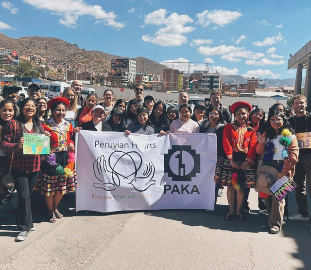 Seventeen Peruvian scholarship recipients standing outdoors holding a PAKA banner against a mountainous town backdrop