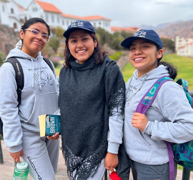 Three Peruvian scholarship recipients smiling on a campus lawn, two in gray Peruvian Hearts hoodies and navy caps with backpacks
