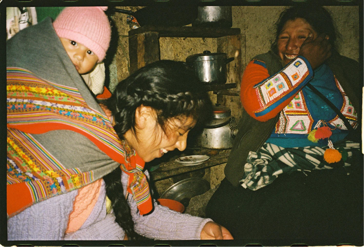 Three people sharing a laugh inside a rustic kitchen, one woman carrying a baby in a colorful woven wrap